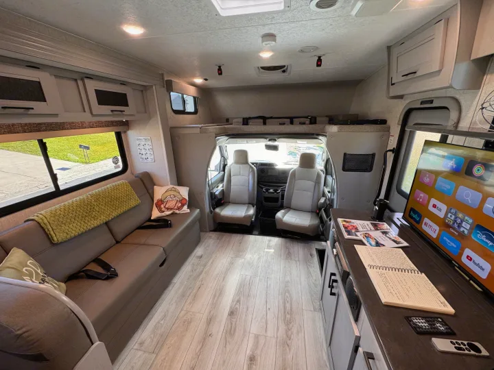 This RV rental's bright interior features wood-look flooring, a comfy sofa, TV entertainment, and front cockpit access, creating a spacious living area.. Forest River Coachmen Freelander 2025