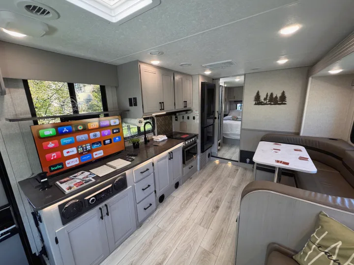 Experience comfort in this spacious RV rental interior, featuring a modern kitchen, TV-equipped living area, and dining nook.. Forest River Coachmen Freelander 2025