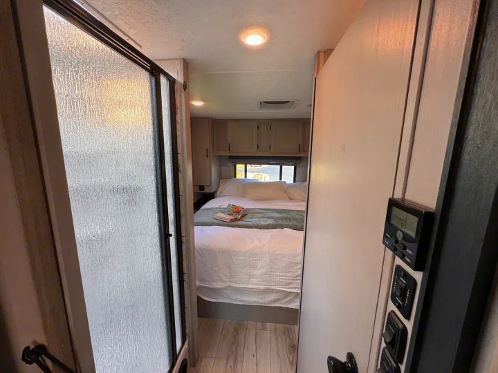 Inside this rv rental: a comfy bedroom area, frosted shower door, and wall-mounted controls.. Forest River Coachmen Freelander 2025