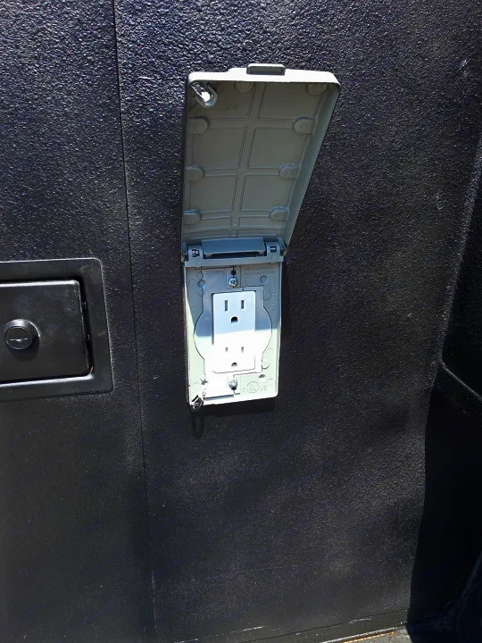 RV rental outdoor weatherproof outlet: A white 3-prong receptacle with an open gray cover on a dark, textured RV exterior.. Custom Build Offroad Trailer 2021
