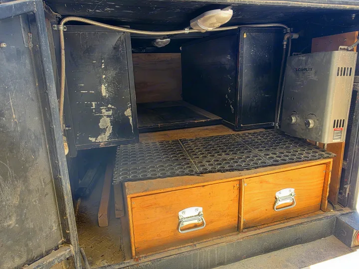 RV rental utility space with a Camplux water heater, black storage cabinets, wooden drawers, and overhead lighting.. Custom Build Offroad Trailer 2021