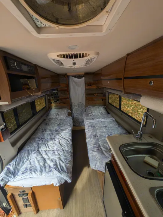 Inside this cozy rv rental: twin beds with patterned comforters, a compact kitchen with sink, wood walls, and overhead storage.. Winnebago Travato 2016