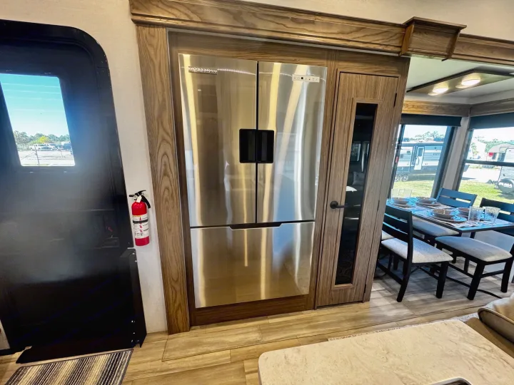 Inside an RV rental: sleek stainless steel refrigerator, fire extinguisher, and dining area with large windows.. Alliance RV Paradigm 2024