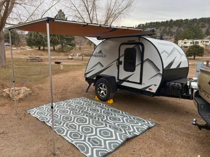 Set up at a serene campsite, this rv rental teardrop trailer features an extended awning and patterned outdoor rug.. Braxton Creek Free Solo 2022