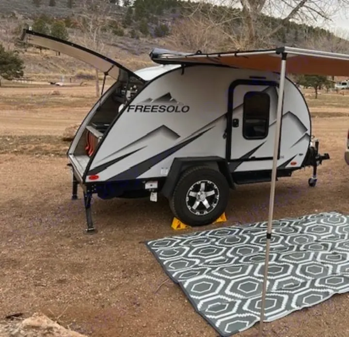 Compact FREESOLO teardrop RV rental on a dirt campsite. Its rear galley is open, and a side awning covers a camping mat.. Braxton Creek Free Solo 2022