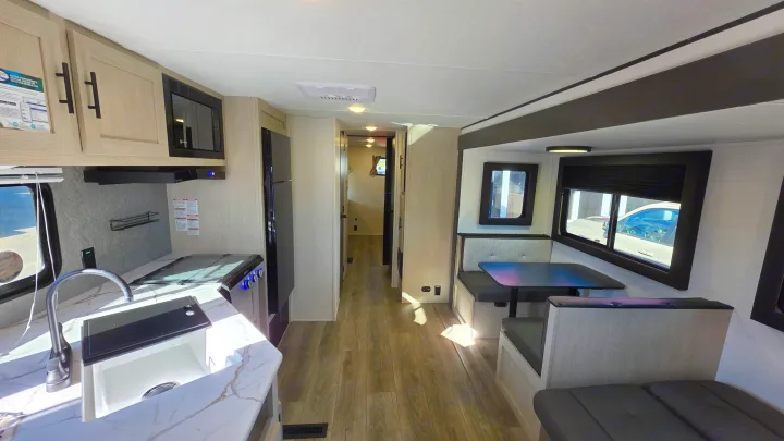 Bright RV rental interior: kitchen with light wood cabinets, sink, microwave, and a dining booth, leading to a hallway.. Forest River Shasta I-5 2025