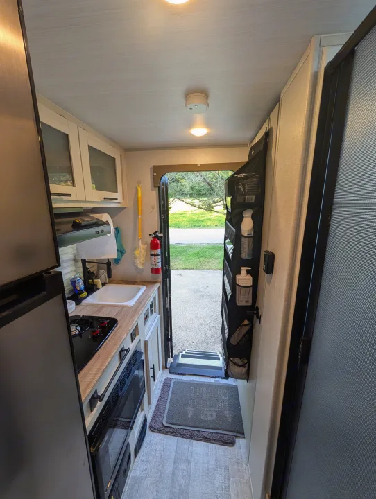 This rv rental shows a compact kitchen with a fridge, sink, and stove, leading to an open entryway with hanging storage.. No Boundaries 16 2024