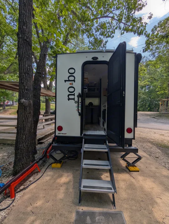 This white "no bo" travel trailer rv rental is set up in a wooded campground, with its door open and steps extended.. No Boundaries 16 2024