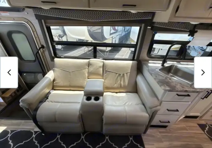 RV rental interior: light beige reclining sofa with cup holders, a large window, and a compact kitchen sink area.. Thor A.C.E. 2021
