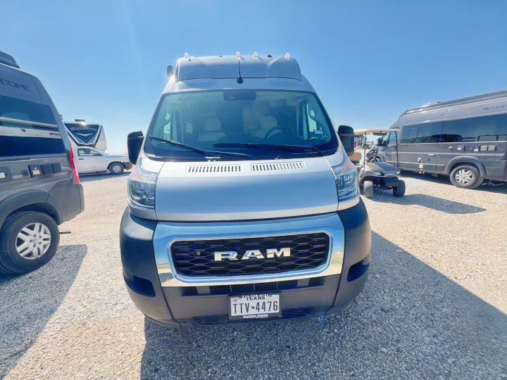 Front view of a silver Ram ProMaster rv rental van parked on a sunny gravel lot, with other RVs and a golf cart visible.. Dodge Ram 1500 2022