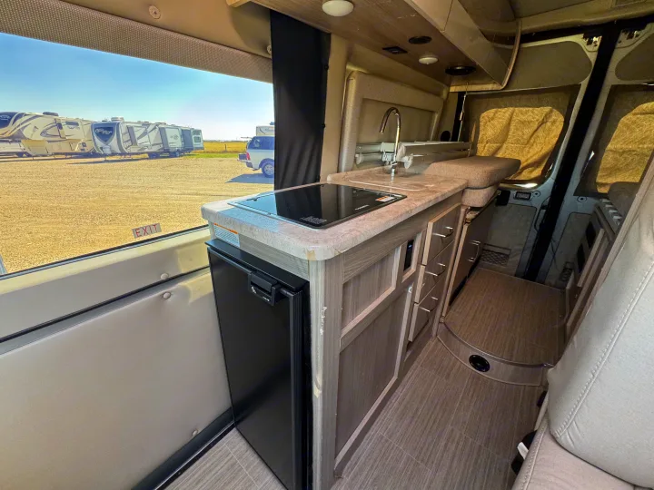 Modern rv rental interior: kitchen with sink, cooktop, and fridge, beside a window revealing parked RVs in an outdoor lot.. Dodge Ram 1500 2022