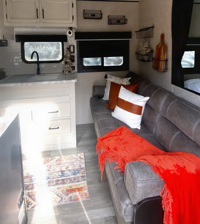 Bright RV rental living space with a grey sofa, orange blanket, and a "Welcome" window sign over the sink in the kitchen area.. Jayco Jay Flight 2023