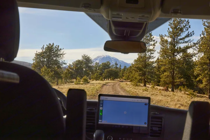 RV rental adventure: Dirt road leads to a majestic snow-capped mountain through a pine forest, navigation active on the screen.. Ford Transit 2023