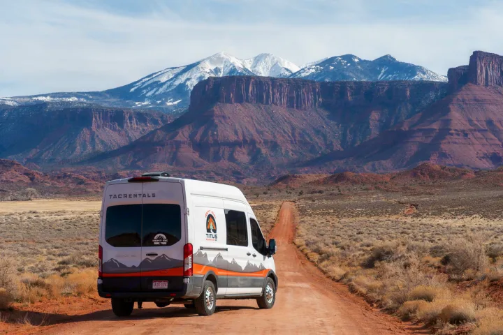 RV rental camper van on a dirt road, with breathtaking red rock mesas and snow-capped mountains beyond.. Ford Transit 2023