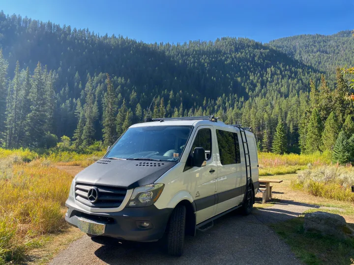 A white Mercedes Sprinter van, an ideal RV rental, parked at a scenic campsite with a lush mountain forest backdrop.. Mercedes-Benz Sprinter 2014