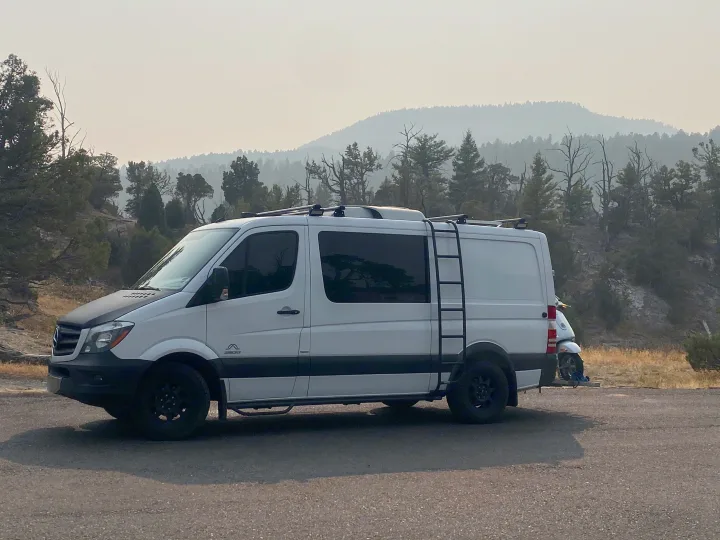A white RV rental camper van, equipped with a ladder and scooter, sits in a hazy, tree-covered landscape.. Mercedes-Benz Sprinter 2014