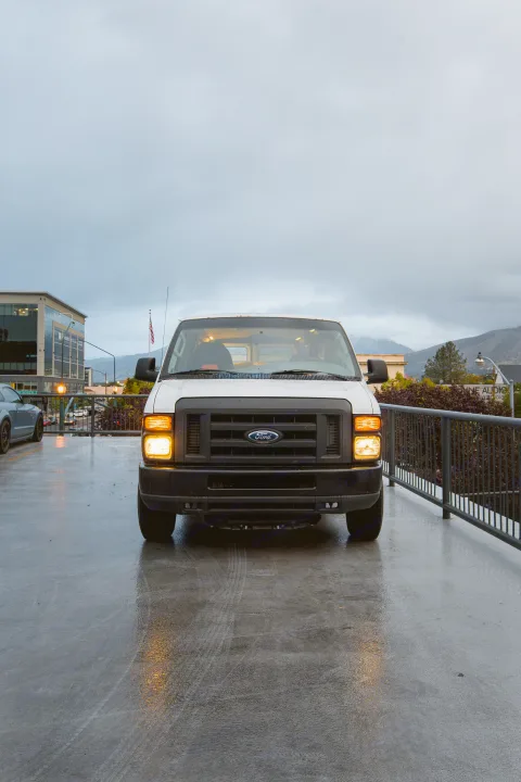This white van, with headlights on, sits on a wet surface. Could be part of an rv rental fleet.. Ford Econoline E250supercab 2008