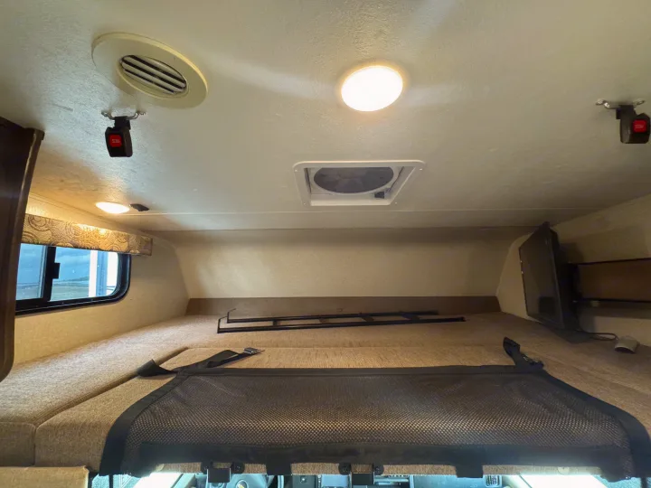Overhead bunk in an RV rental, featuring a safety net, window, TV, AC vent, and ceiling fan for a comfortable stay.. Forest River Coachmen Freelander 2019