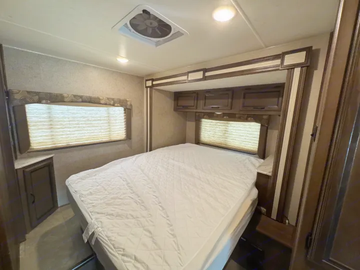 This RV rental bedroom features a cozy bed with white bedding, overhead storage, twin windows with blinds, a ceiling vent, and lights.. Forest River Coachmen Freelander 2019