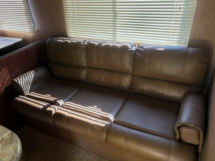 Comfortable brown leather couch in an RV rental, with sunlight streaming through window blinds, offering cozy seating for travelers.. Dutchmen Coleman Lantern 2018