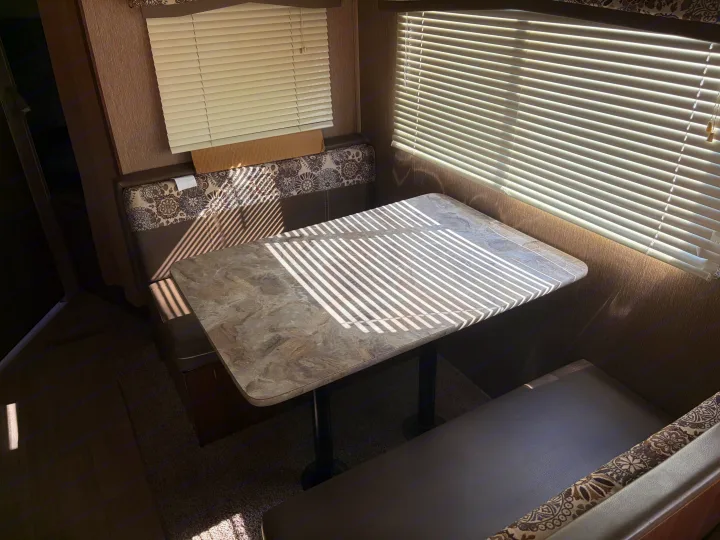 Cozy RV rental dinette bathed in sunlight from closed blinds, creating striped shadows on the table and patterned benches.. Dutchmen Coleman Lantern 2018