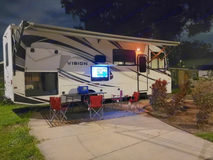 An inviting RV rental setup for an evening outdoors, featuring an extended awning, outdoor TV, kitchen, and chairs.. Entegra Coach Vision 2019