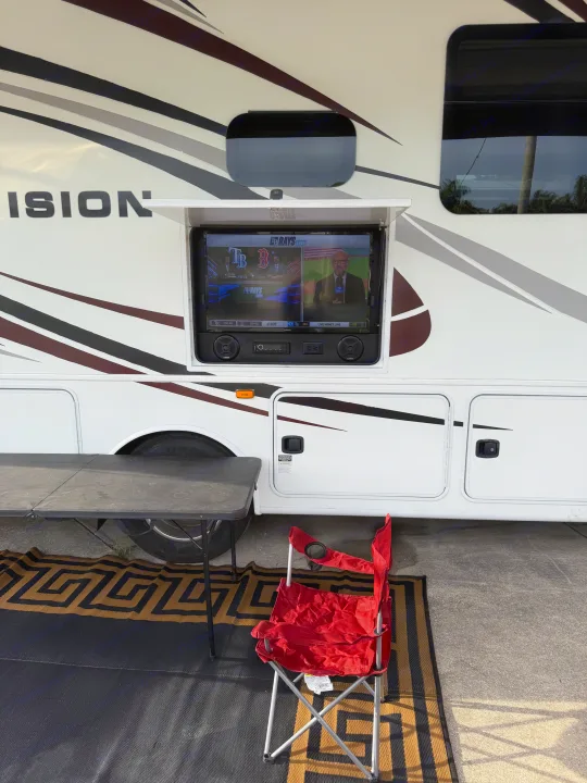 RV rental with outdoor TV showing a live baseball game, complemented by a folding table, chair, and mat for outdoor enjoyment.. Entegra Coach Vision 2019