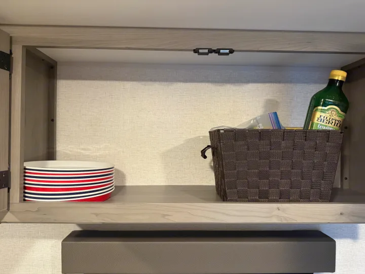 Inside an RV rental cabinet: striped plates, a woven basket holding Filippo Berio olive oil, and magnetic latches.. Keystone RV Hideout Sport 2025