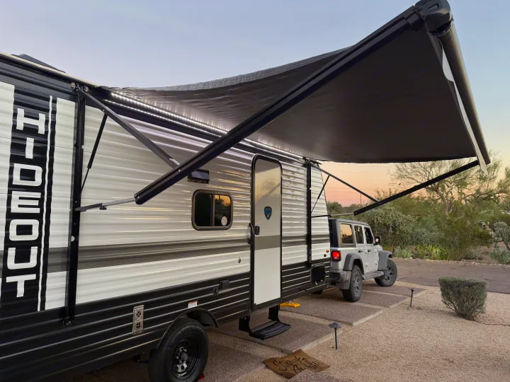 An RV rental travel trailer with an extended, illuminated awning, parked with a Jeep at a desert campsite at dusk.. Keystone RV Hideout Sport 2025