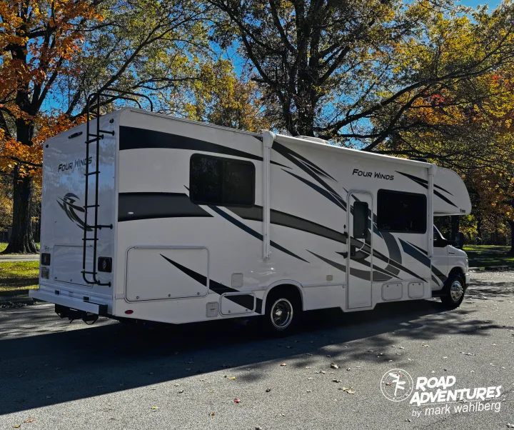 A white "Four Winds" rv rental with black graphics is parked by sunny autumn trees, ready for a fall road trip adventure.. Thor Motor Coach Four Winds 28Z 2024