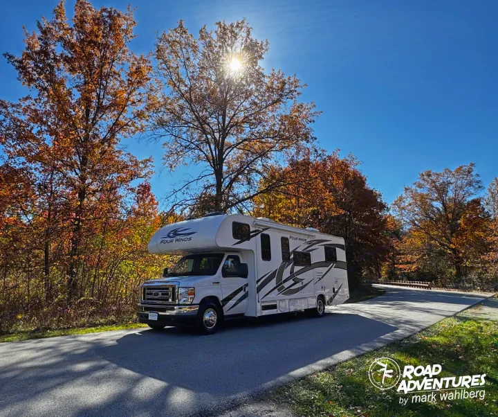 A white Four Winds RV rental drives on a paved road through trees with bright autumn leaves under a sunny blue sky.. Thor Motor Coach Four Winds 28Z 2024