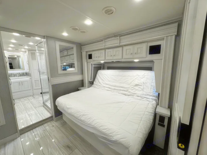 Modern RV rental bedroom features a large white bed, overhead cabinets, and an en-suite bathroom with a vanity visible.. Holiday Rambler Armada 2022