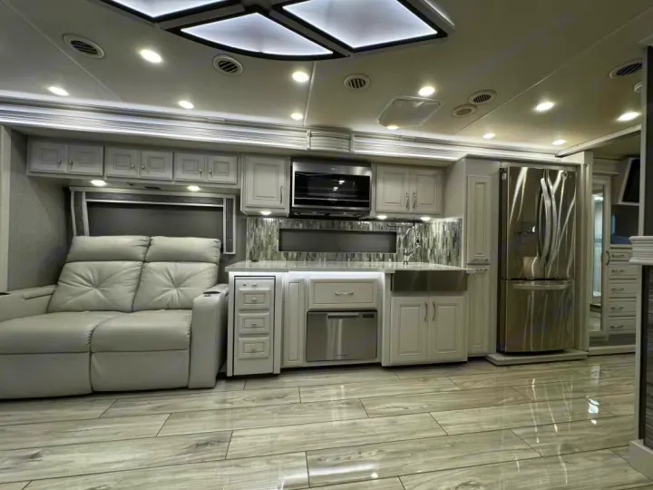 Modern RV rental interior: A full kitchen with stainless appliances, light cabinetry, a tiled backsplash, and a plush gray reclining sofa.. Holiday Rambler Armada 2022
