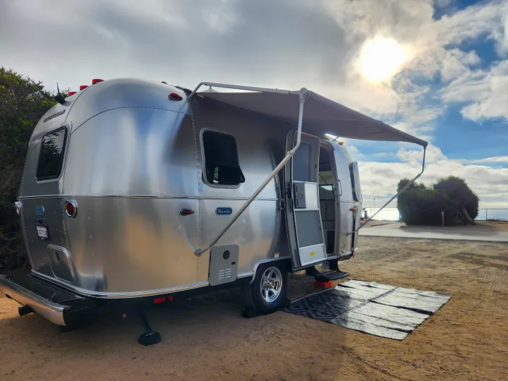Experience an RV rental with this Airstream Bambi parked seaside, awning out under a sunny, cloudy sky.. Airstream Bambi 2023