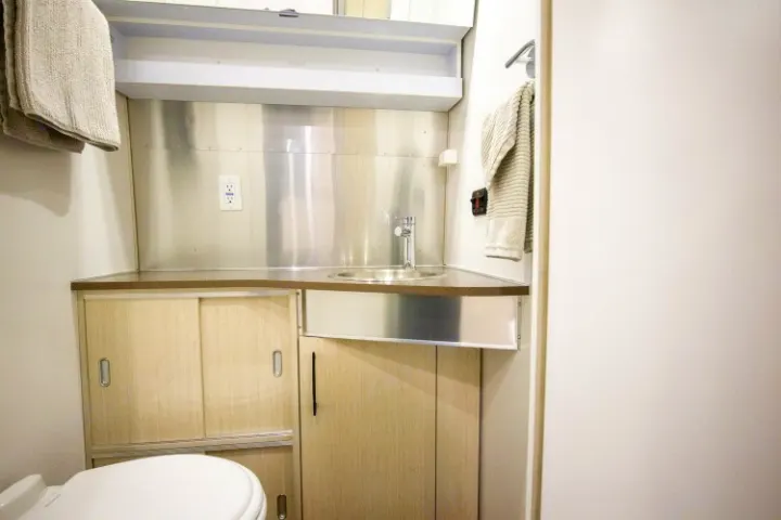 Compact rv rental bathroom vanity with sink, toilet, wood cabinets, & stainless steel backsplash. Towels hang on a rack.. Airstream International 2020