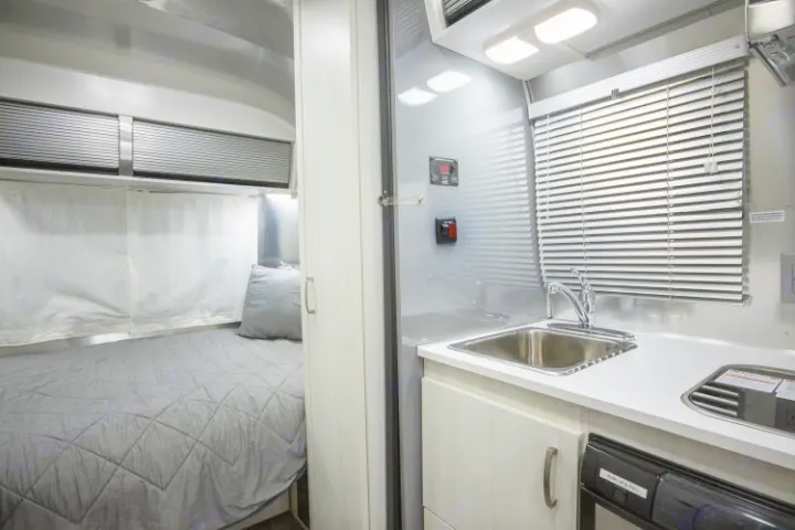 This compact RV rental interior features a cozy bedroom with a quilted grey bed and an efficient kitchen area with a sink and stovetop.. Airstream Sport 2019