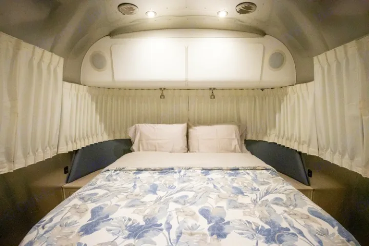 Inside an RV rental: a cozy bedroom with a floral bedspread, white curtains, reading lights, and overhead storage.. Airstream Globetrotter 2019