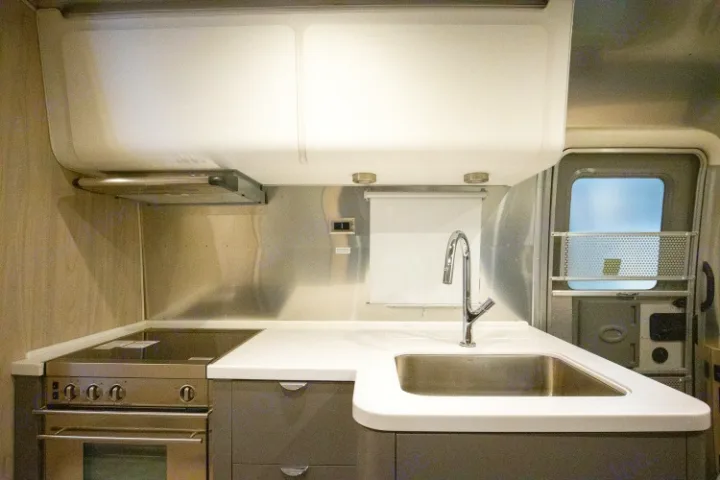 View of a modern rv rental kitchen, featuring a stainless steel oven, cooktop, sink, faucet, and white countertops.. Airstream Globetrotter 2019