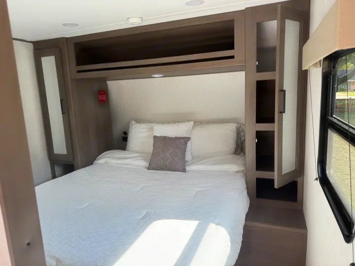 An RV rental bedroom with a neatly made white bed, wooden storage cabinets, an overhead light, and a window.. Transcend 26BHX 2025