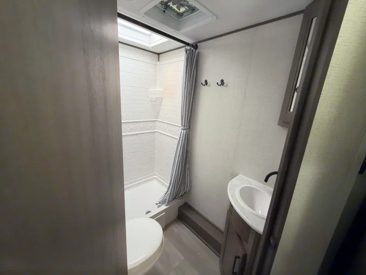 Functional RV rental bathroom with a white brick-patterned shower, striped curtain, toilet, sink, & dark vanity.. Transcend 26BHX 2025