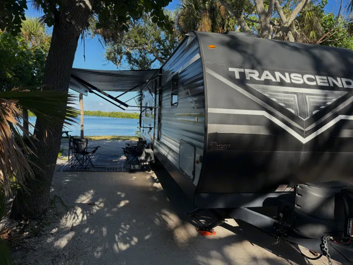 Scenic waterfront rv rental setup: a travel trailer with an extended awning, chairs, and rug, shaded by trees overlooking the water.. Grand Design Transcend Xplor 2022