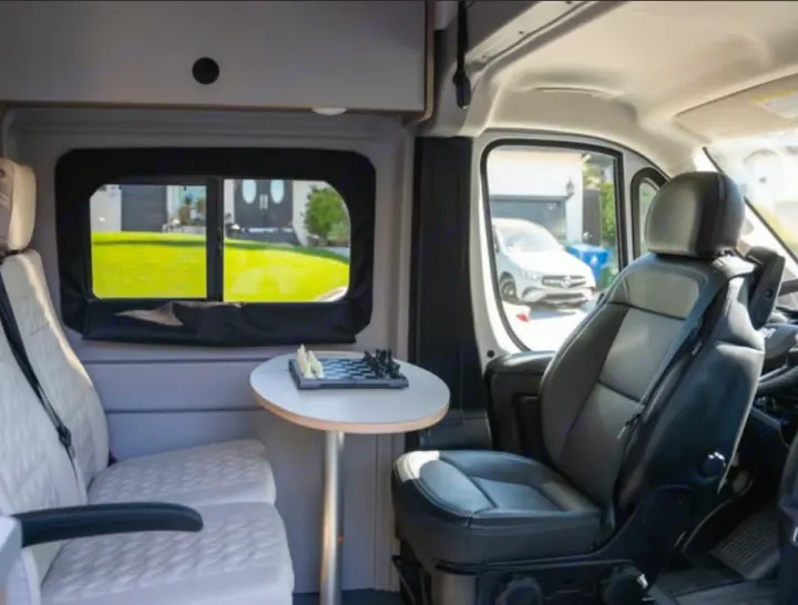 Inside a spacious RV rental: light seats, table with chess set, driver's seat visible. View of a house outside.. Winnebago Solis 2025