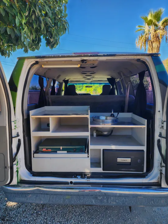 Rear view of an RV rental camper van's custom kitchen: sink, pull-out stove, Dometic fridge, and storage shelves.. Escape Trailer Industries E150 XLT 2011