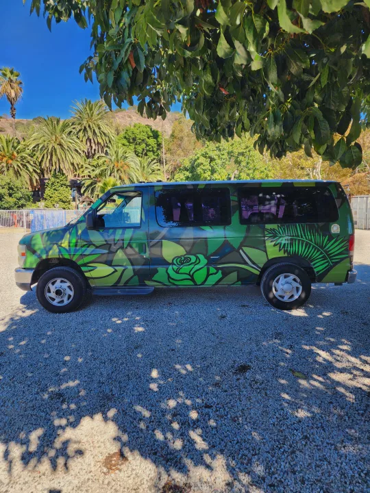 This vibrant green RV rental van, custom-painted with leaf and rose designs, is parked on gravel under a tree on a sunny day.. Escape Trailer Industries E150 XLT 2011
