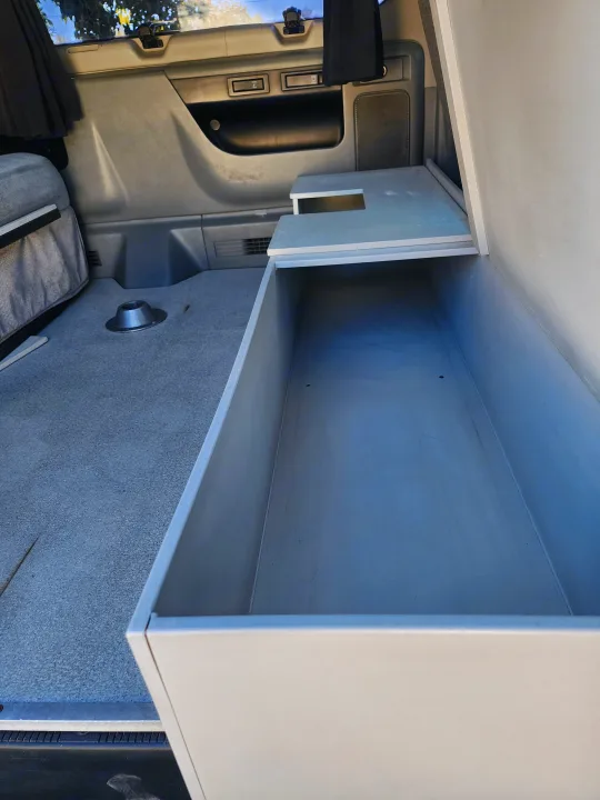 Inside this **rv rental**, a spacious built-in storage box is open, revealing its deep interior alongside a carpeted floor with a table mount and vehicle side panels.. Escape Trailer Industries E150 XLT 2011