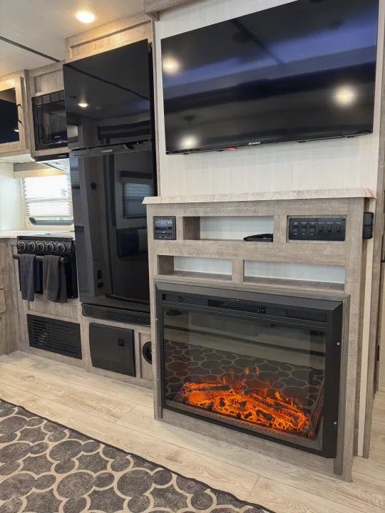 RV rental living space: a flat-screen TV above an electric fireplace, controls, and kitchen area with oven, microwave, and fridge.. Forest River Surveyor 2023