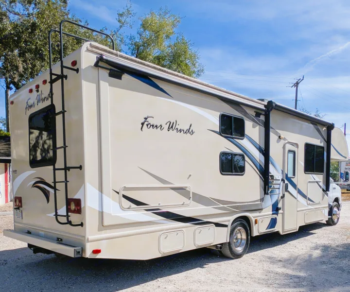 Beige Four Winds RV rental, a Class C motorhome, parked on gravel. Features include a rear ladder, side awning, and storage bays.. Thor Four Winds 2017