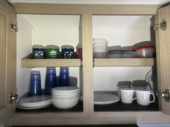 Inside an RV rental kitchen cabinet: camping mugs, blue cups, plates, bowls, and storage containers, all neatly stored for your trip.. Forester LE 2026