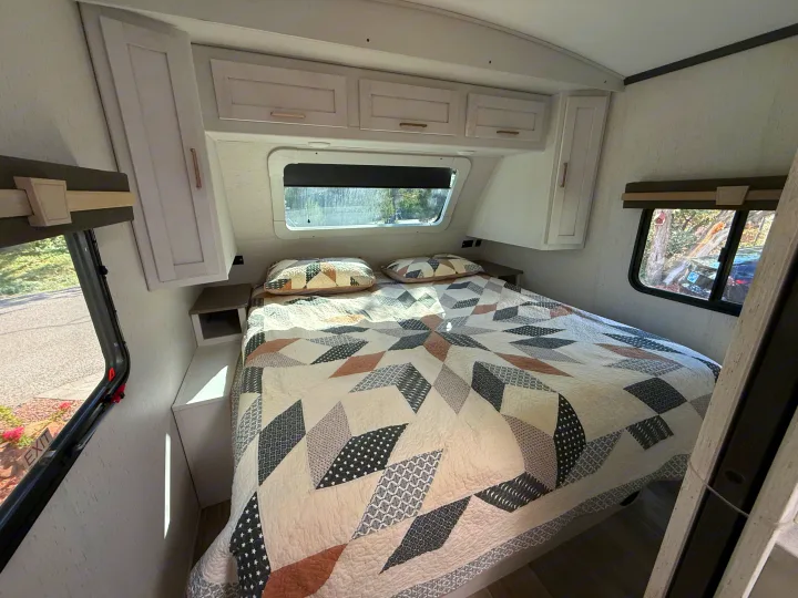 Experience comfort in this RV rental bedroom: a cozy bed with a geometric quilt, overhead cabinets, and bright windows.. Astoria Astoria Trailer 2022