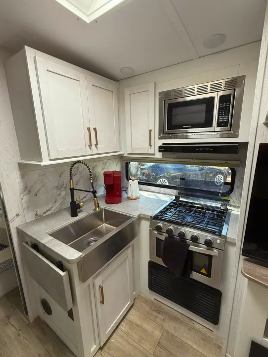 Modern RV rental kitchen featuring a farmhouse sink, stove, microwave, and light-colored cabinets with brass handles.. Astoria Astoria Trailer 2022